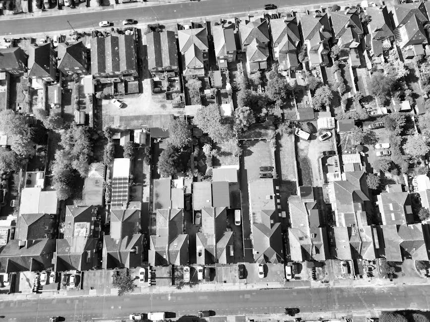 A street view in Queens Park with residential brick and concrete buildings lining both sides of the road. The street is narrow, with parked cars along the sidewalks and a silver Audi driving towards t