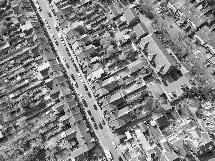 Aerial view of a residential street showing a row of houses with pitched roofs and small backyards, parked cars lining both sides of the road, and trees scattered along the pavement. The street appear